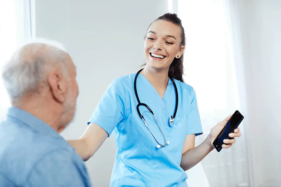 Eine Krankenschwester steht mit einem Telefon vor einem älteren Herren in einem Pflegezentrum.